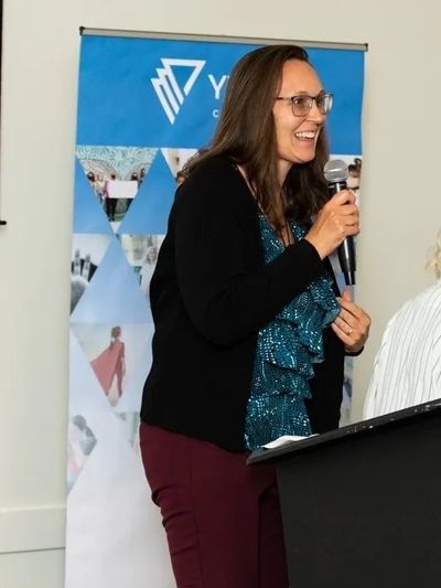 A woman standing at a podium holding a microphone and smiling in front of a blue banner.