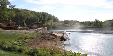 Construction equipment near a water body surrounded by trees on a sunny day.