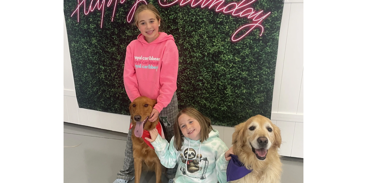 Two girls smiling with their dogs in front of a "Happy Birthday" sign.