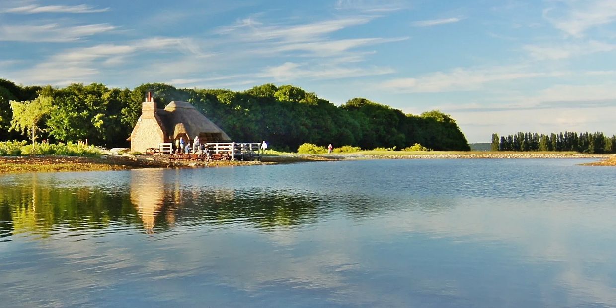 Thatched cottage by a calm lake with lush green trees and a blue sky.
