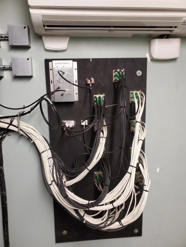Organized black and white cables connected to a black panel under an air conditioner.
