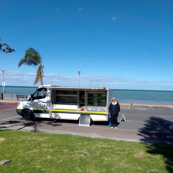 A food truck parked by the seaside with a woman standing beside it on a sunny day.