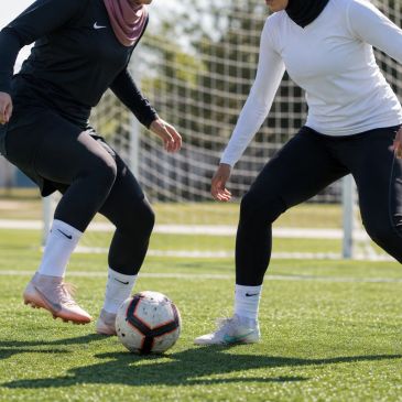 Two women playing soccer wearing hijabs and sportswear.