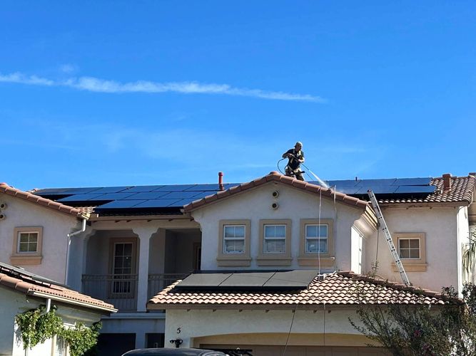 Worker cleaning solar panels on a sunny roof with a pressure washer.