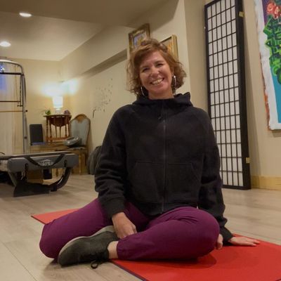 A woman in purple pants sitting on a red yoga mat indoors, smiling.  Jennifer Carmack at  Phoenix Rising Pilates in Fort Collins, Colorado