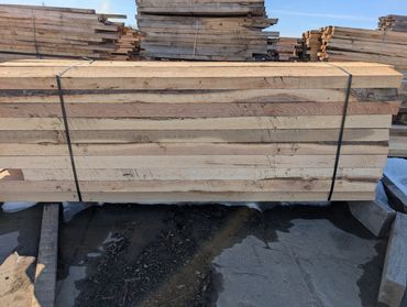 Stack of rough-cut lumber secured with black straps outdoors.