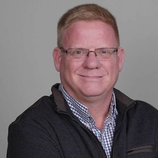 Smiling man with glasses in a checkered shirt and dark jacket.