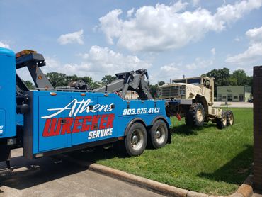 Blue Athens Wrecker Service truck towing a large beige military truck on a grassy area.