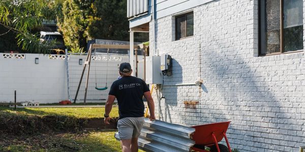 Man carrying metal sheets near a white brick house with a red wheelbarrow.