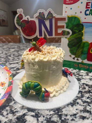 A white birthday cake decorated with "The Very Hungry Caterpillar" theme and a "ONE" topper.