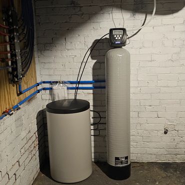 Water softener and filtration system installed in a basement corner with white brick walls.