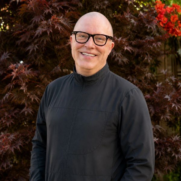 Smiling man with glasses in black jacket outdoors.