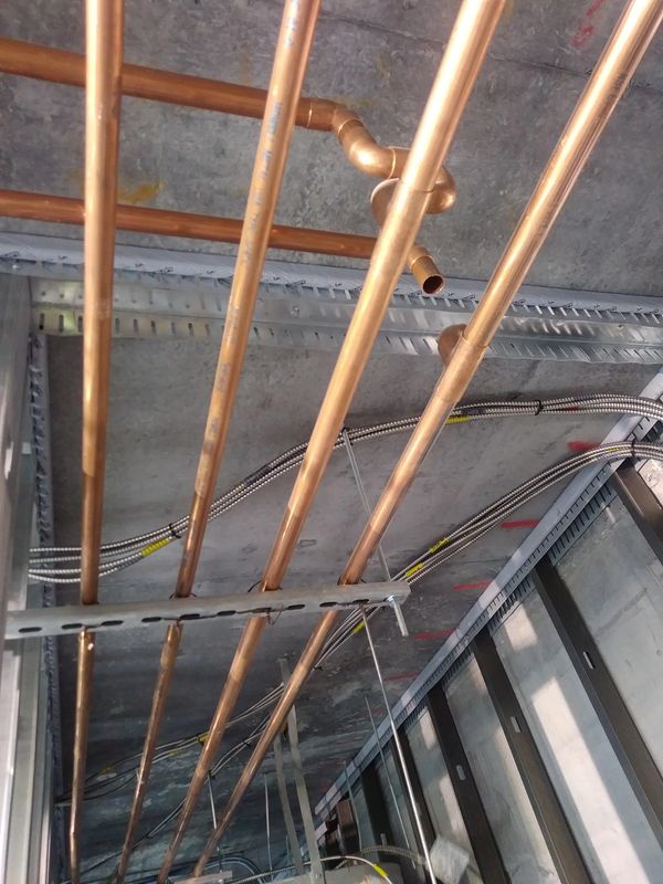 Copper pipes and metal conduits installed on a concrete ceiling in a building under construction.