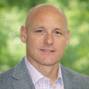 Portrait of a bald man in a gray blazer and checkered shirt.