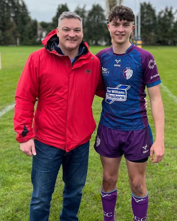 Two men posing on a grassy sports field, one in a red jacket and the other in a rugby uniform.