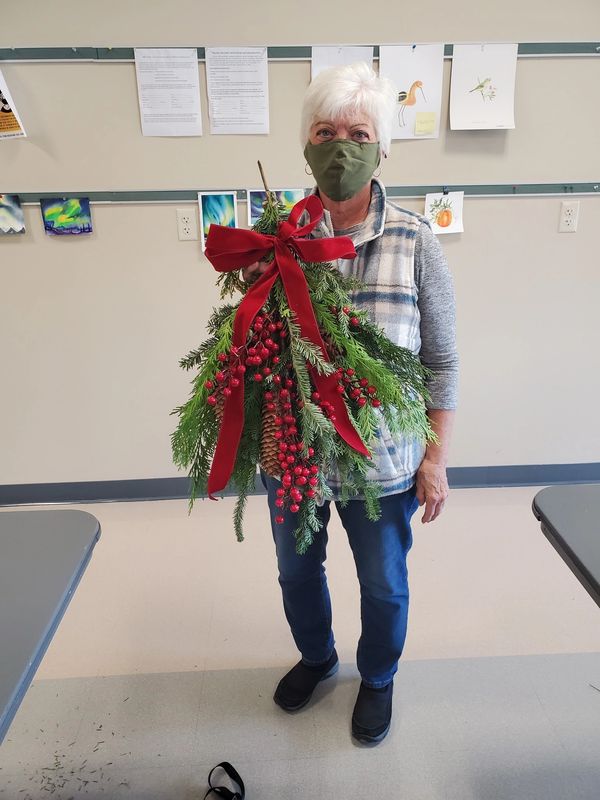 Member poses in green face mask holding a large evergreen swag with berries & bow at ARC