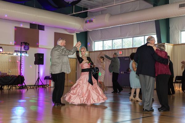 4 couples dance in fancy dresses & suites - woman in foreground twirls in pink ruffle dress