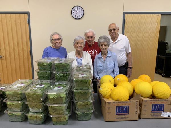 Group of volunteers stand behind table full of spinach containers & squash at food item pick up
