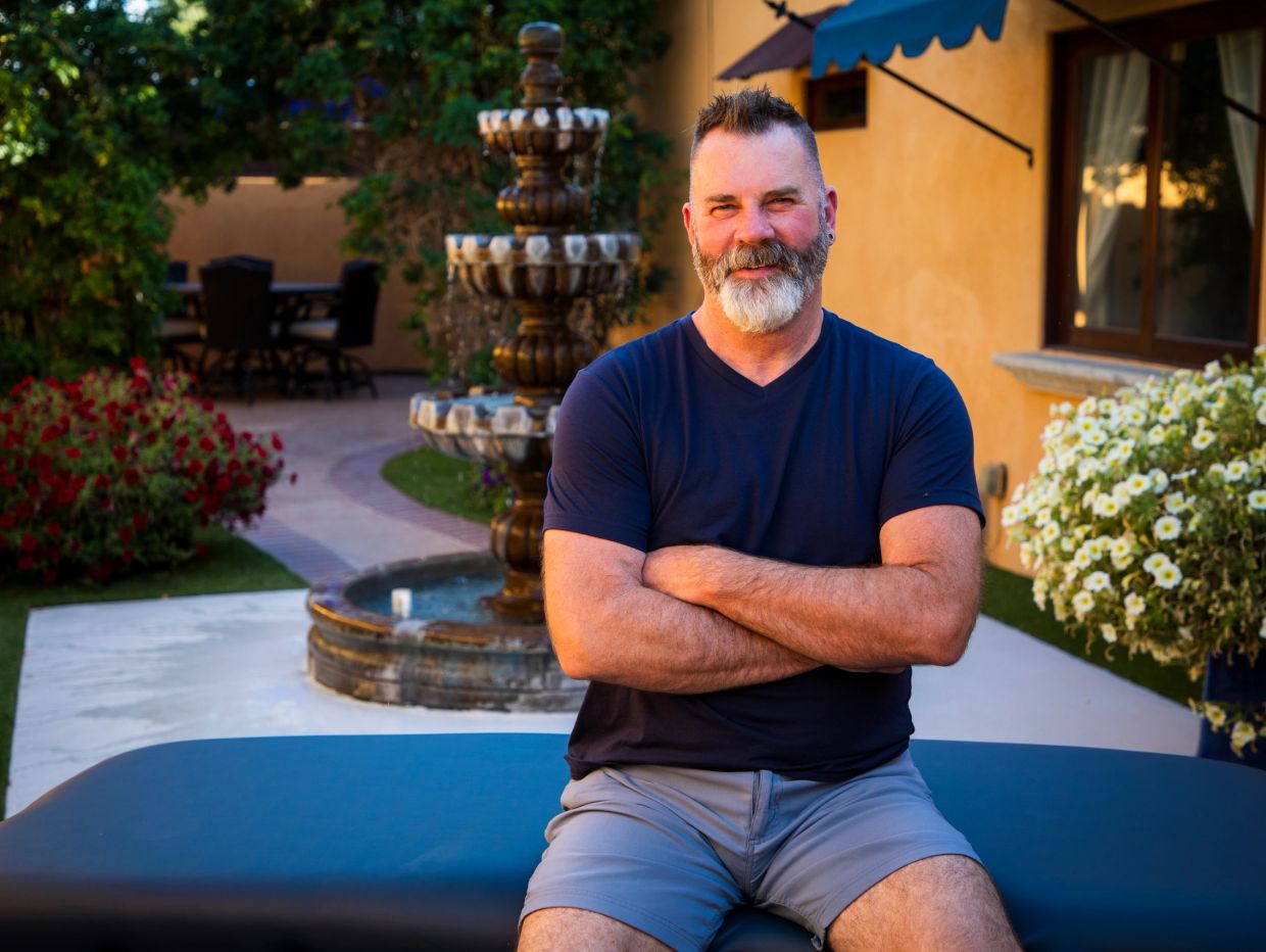 Smiling man with a beard sitting outdoors near a fountain and flowers.