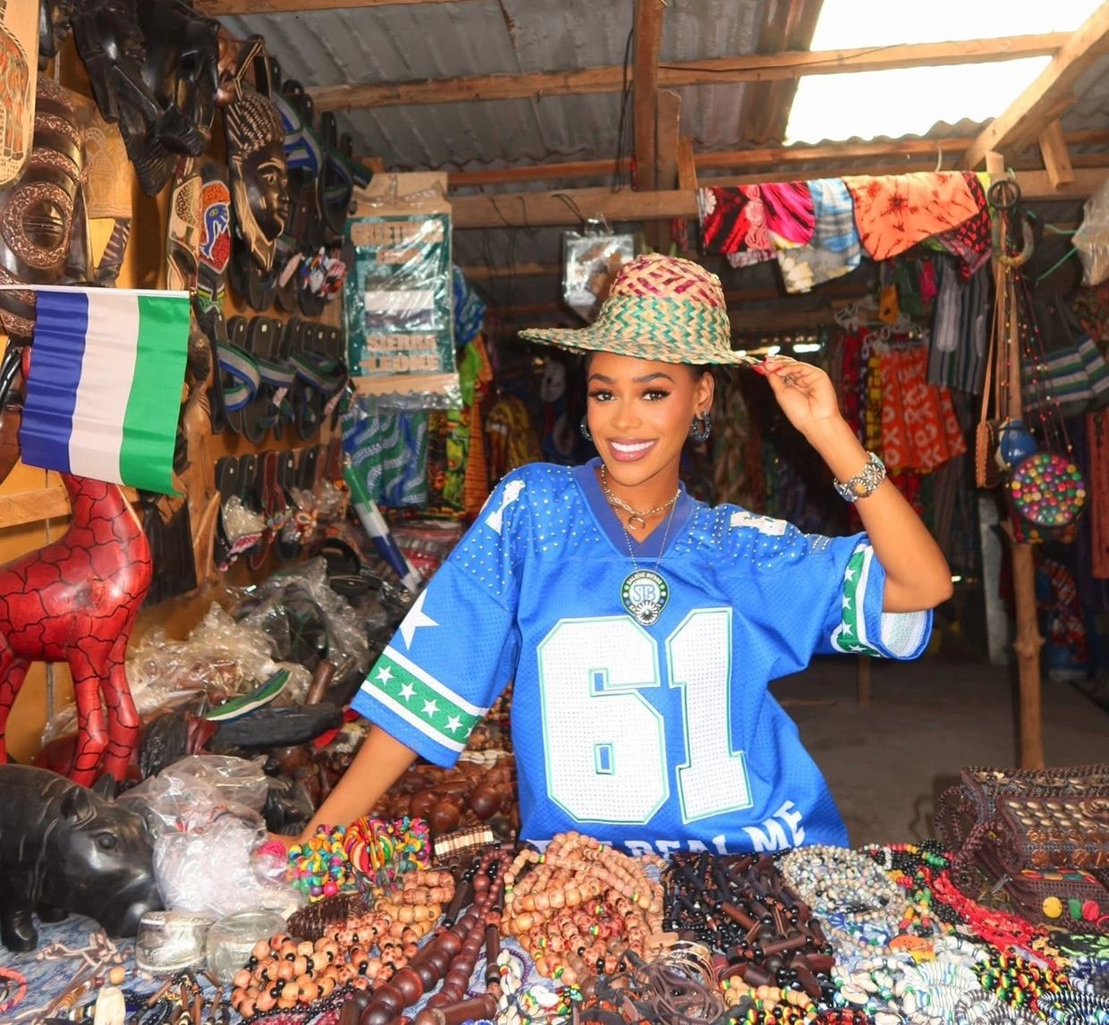 Woman in blue jersey and woven hat selling crafts at a colorful market stall.