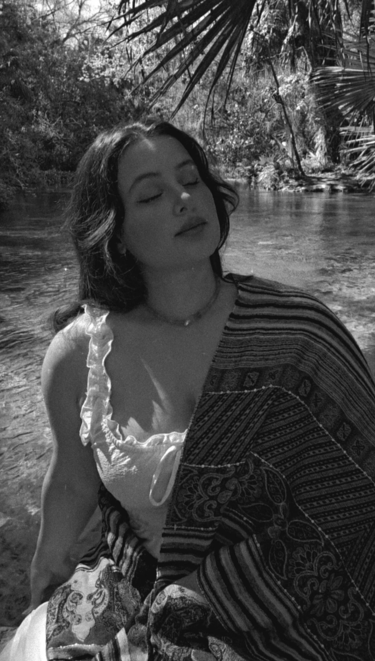 Woman with closed eyes wrapped in patterned fabric by a serene river.