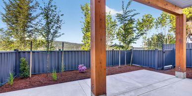 Modern backyard patio with wooden posts and young trees along a blue fence.
