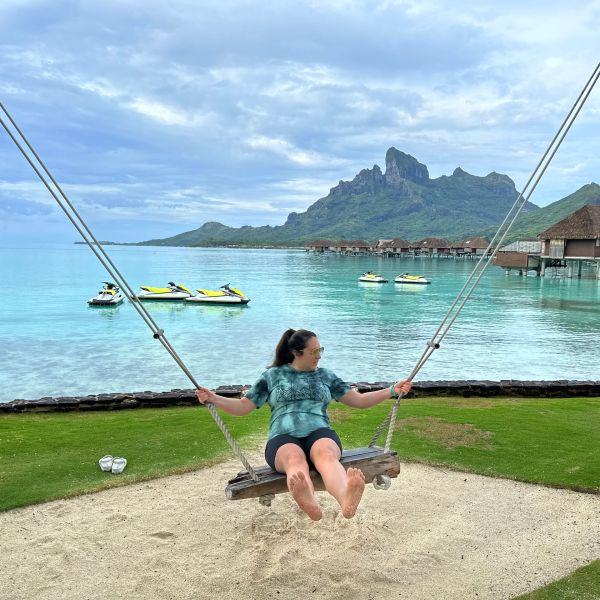 Woman on a swing by the tropical sea with jet skis and mountains.
