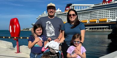 Family of five posing near a dock with a cruise ship behind them.