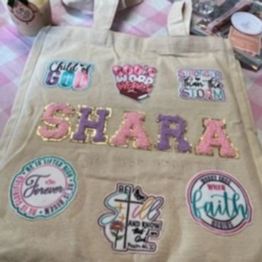 A beige tote bag decorated with colorful faith-inspired patches and the name 'SHARA'.