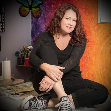 Felisha Bustos Herstrory President sitting in front of a rainbow painted wall and art supplies