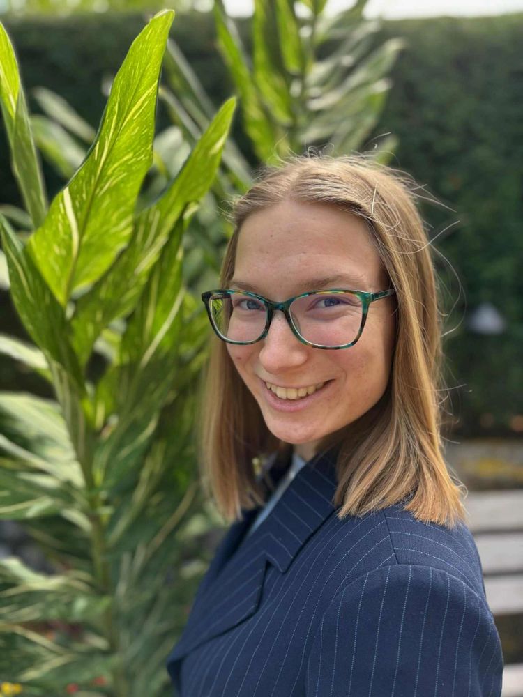 Young woman with glasses smiling outdoors near large green plants.