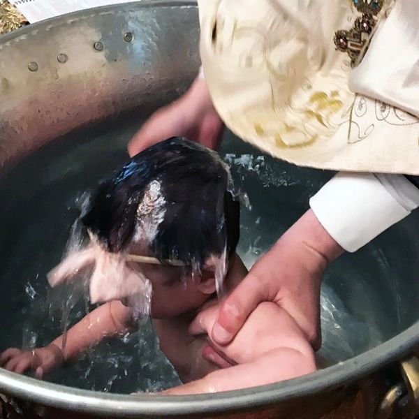 A baby being baptized with water by a priest in a metal basin.