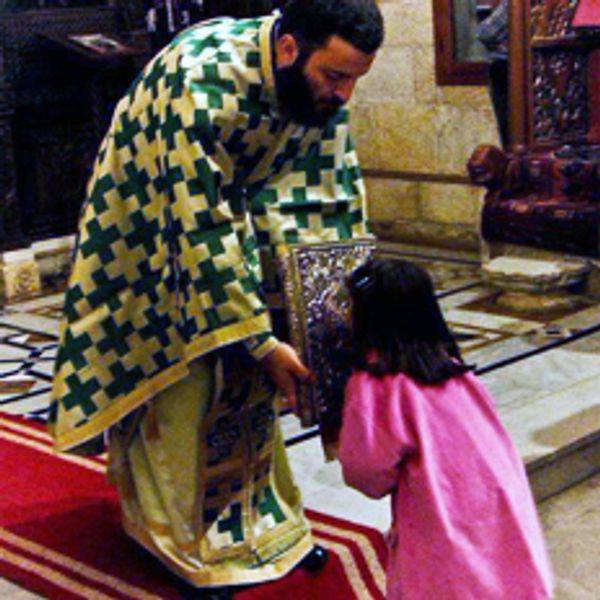 A priest in ceremonial robes presents a religious book to a bowing child in pink.