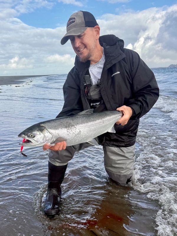 Jayson kneeling in the surf, looking down at a salmon with a pink lure.