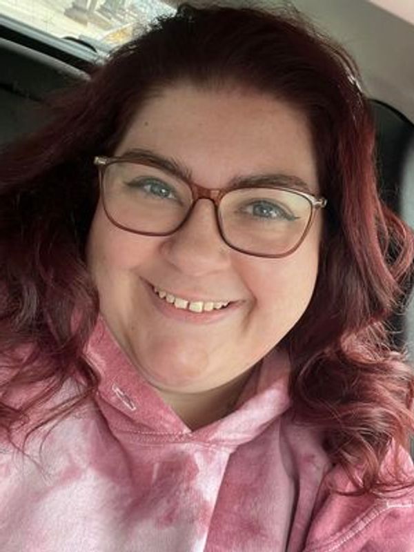 Smiling woman with glasses and curly hair wearing a pink hoodie.