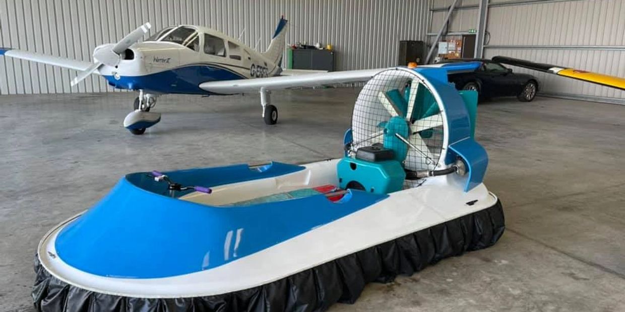 Blue and white hovercraft parked inside a hangar with a small airplane in the background.