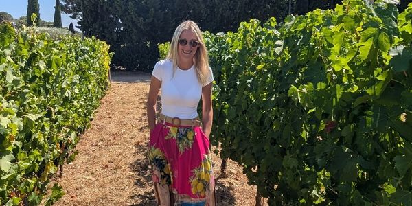 Woman in floral skirt stands in vineyard rows.