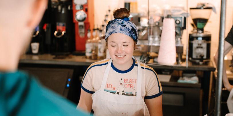 Smiling barista ringing up a doughnut order, with coffee equipment in the background.