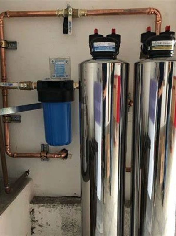 A whole-home water filtration system is installed along a copper plumbing line. The setup includes two tall stainless-steel filtration tanks positioned side by side, paired with a blue pre-filter housing mounted on the wall. Shutoff valves and precision copper piping connect each component, creating a clean, organized installation. The configuration is designed to remove sediment, chemicals, and impurities before water enters the home’s plumbing system, improving overall water quality and protecting fixtures and appliances.