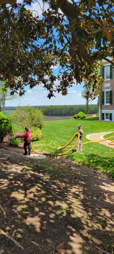 Two workers using large hoses in a landscaped garden near a brick building.