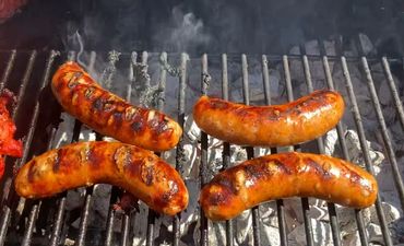 Four sausages grilling over charcoal with smoke rising.