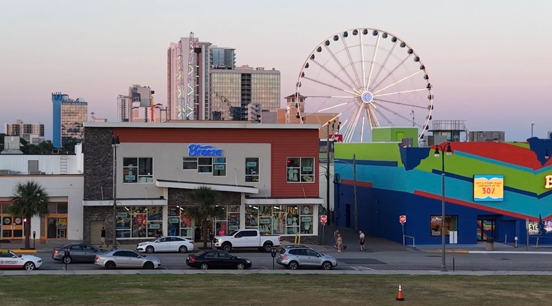 Cityscape with a Ferris wheel, colorful buildings, and cars on the street at dusk.