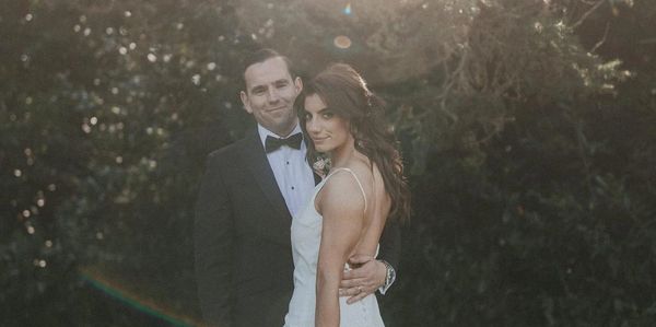 Elegant bride and groom posing outdoors with soft sunlight and floral bouquet.