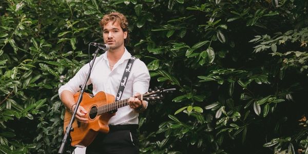 A man plays acoustic guitar and sings in front of lush green foliage.