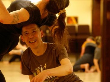 A man laughs as he balances a person leaning back on his head.