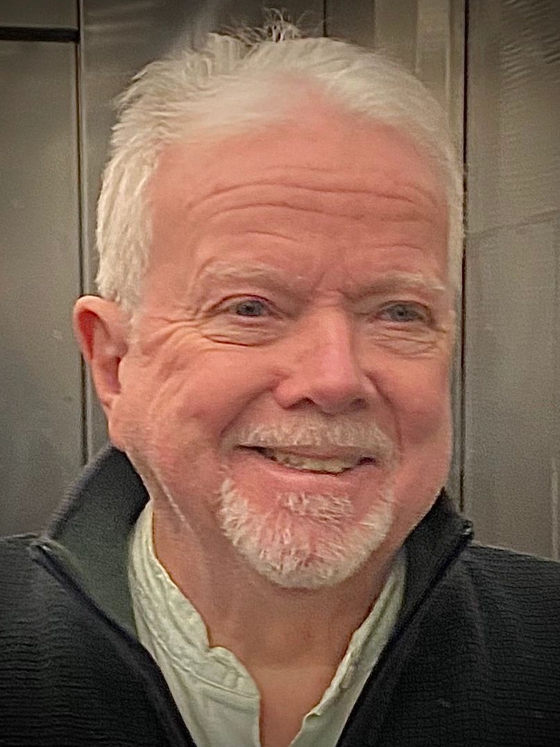 Smiling elderly man with white hair and beard, wearing a black jacket.