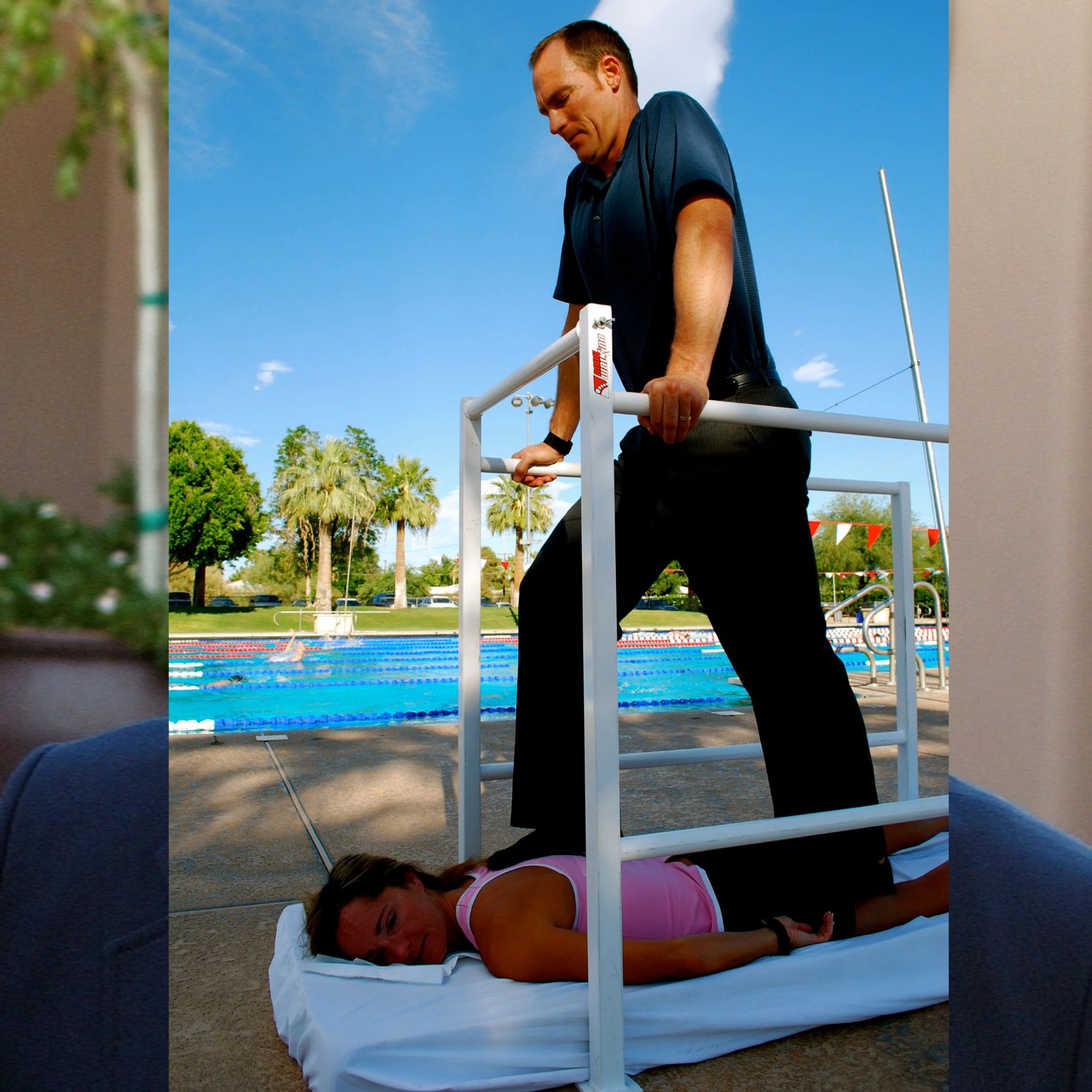 A man performs a back massage using his feet on a woman lying on a mat by the pool.