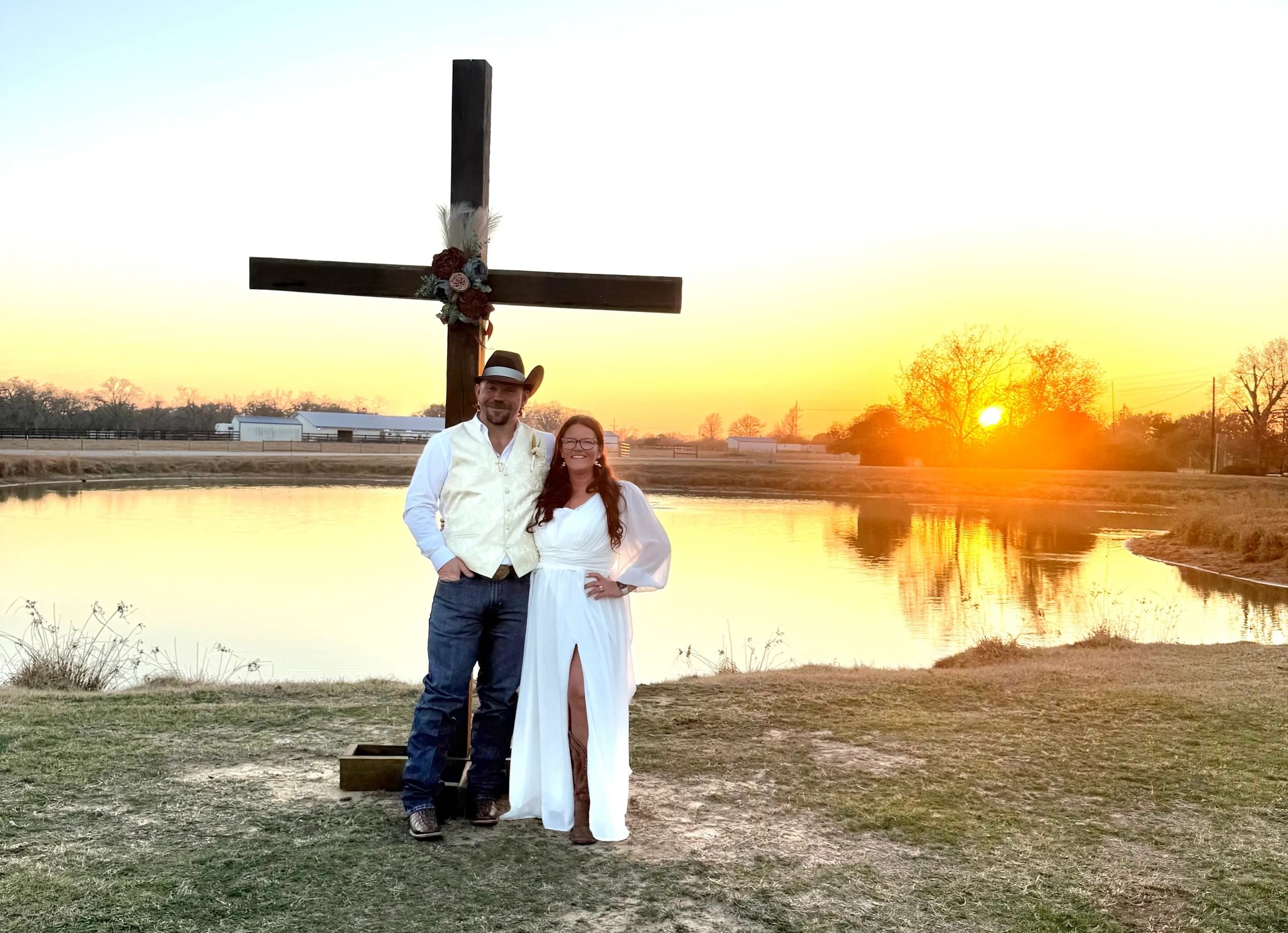 A couple poses by a decorated cross at sunset near a pond.