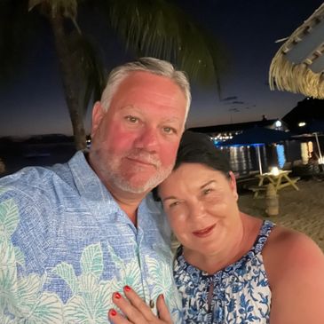 Couple smiling together on a beach at night with palm trees and water in the background.