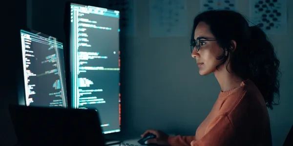 Woman coding on dual monitors in dark room.
